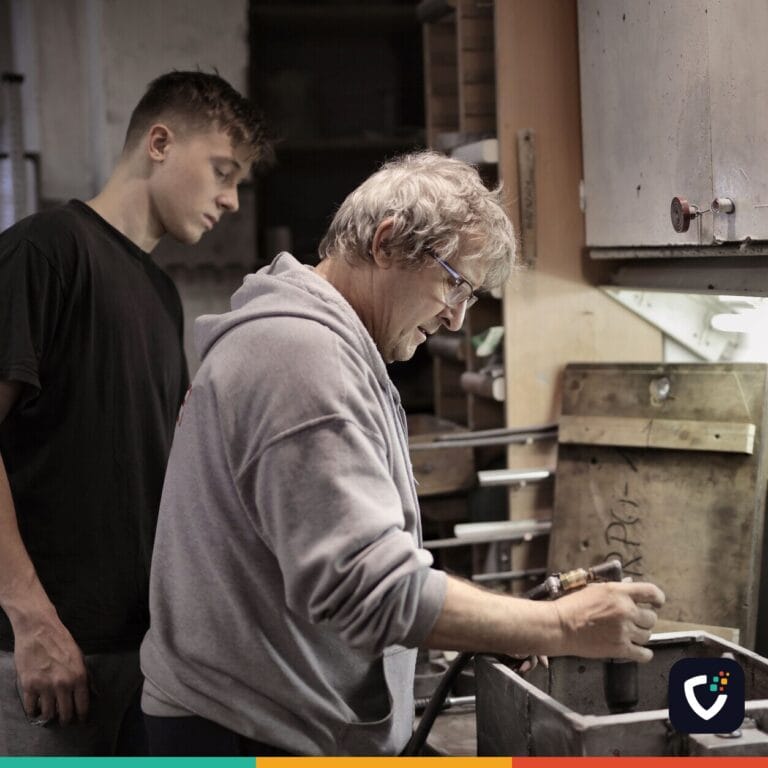 Side view of senior worker in eyeglasses and trainee handling detail on workbench in workshop