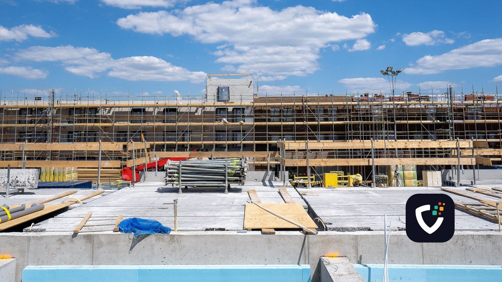 A building under construction, its facade covered in scaffolding, with timber falsework and stacked scaffold tubes on the slab in front of it.