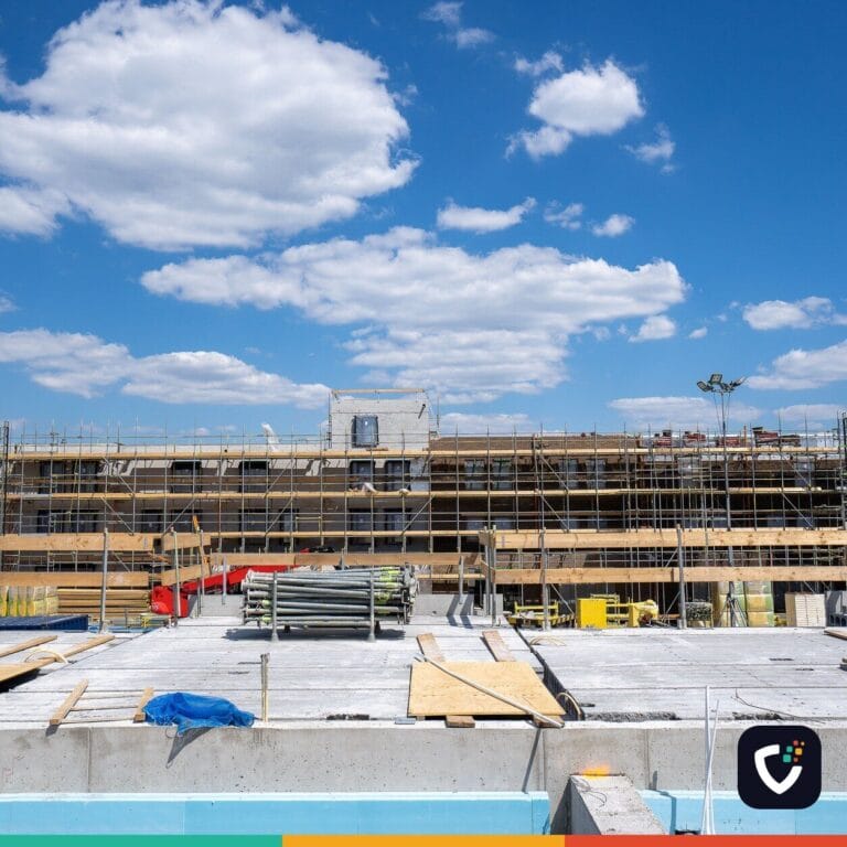 A building under construction, its facade covered in scaffolding, with timber falsework and stacked scaffold tubes on the slab in front of it.
