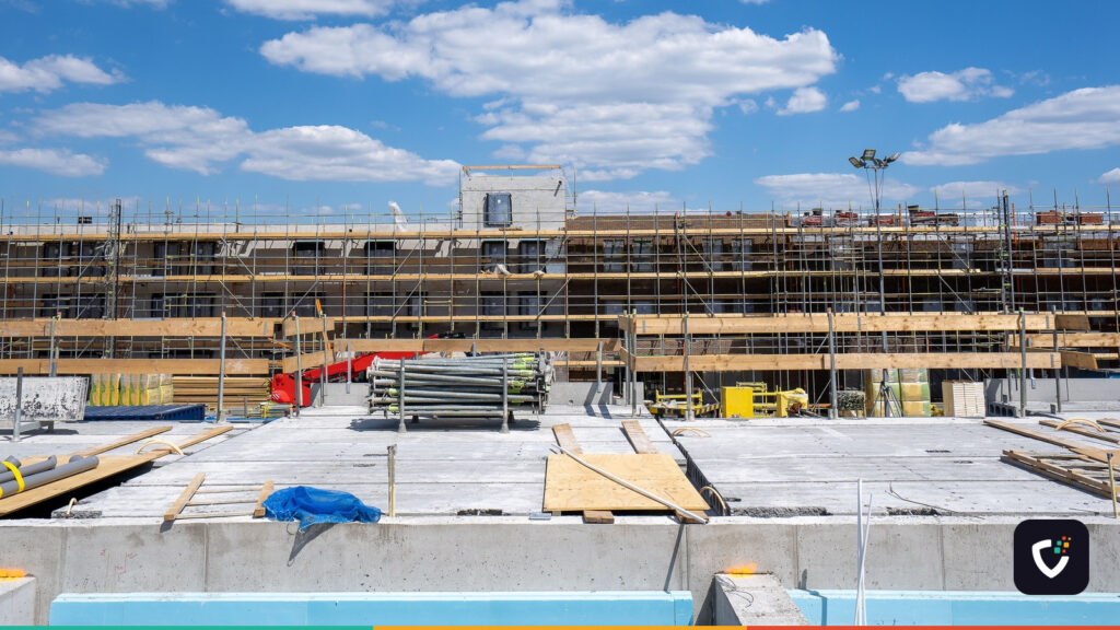 A building under construction, its facade covered in scaffolding, with timber falsework and stacked scaffold tubes on the slab in front of it.