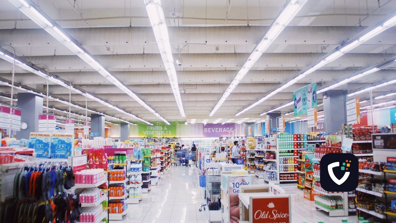 A wide supermarket aisle with lit ceiling strips, well stocked shelves and a few shoppers in the distance.