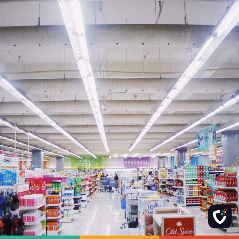 A wide supermarket aisle with lit ceiling strips, well stocked shelves and a few shoppers in the distance.