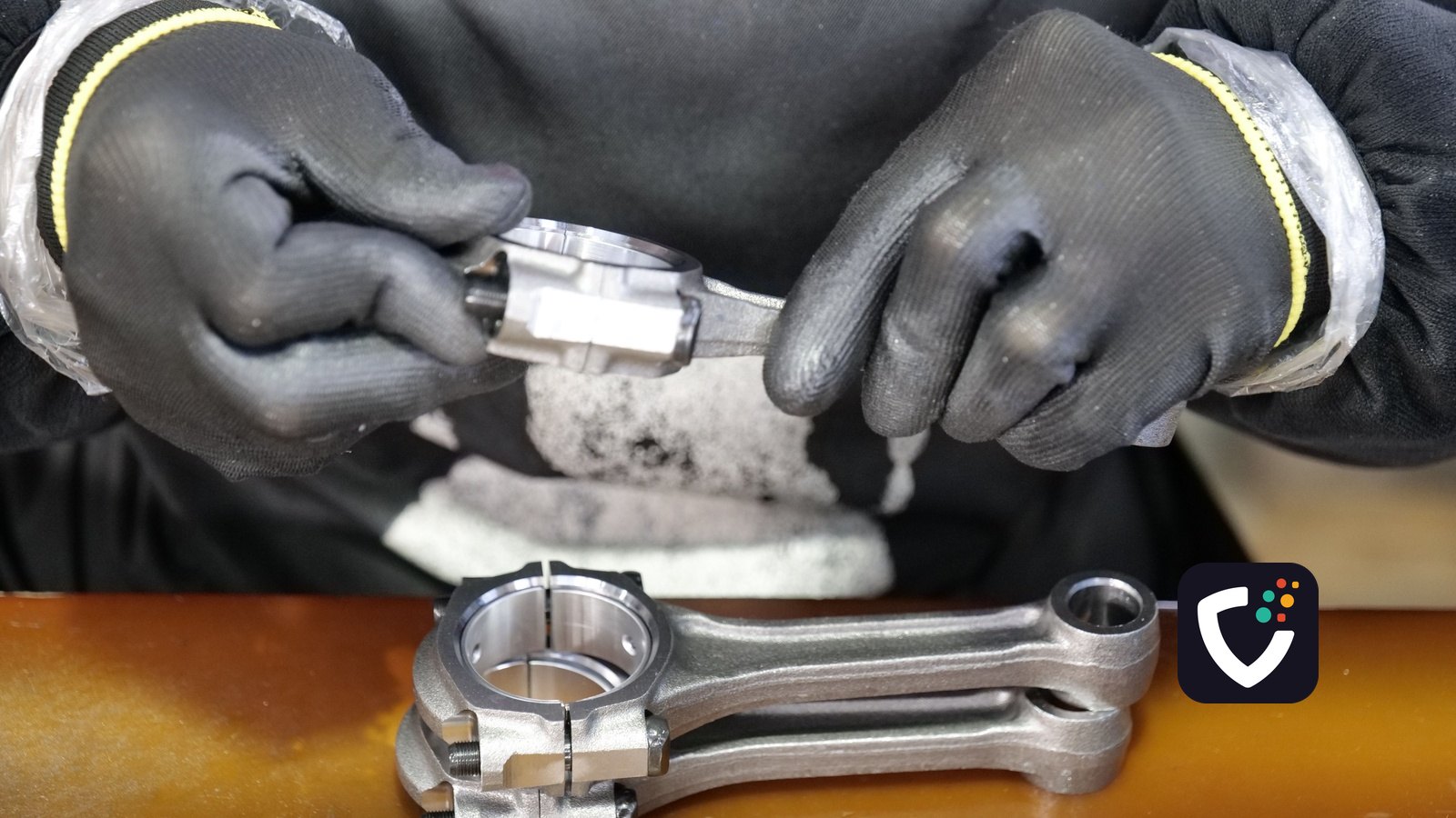 A technician wearing grip gloves checking a connecting rod bearing shell at a workbench, with a second assembled rod resting in front of him.