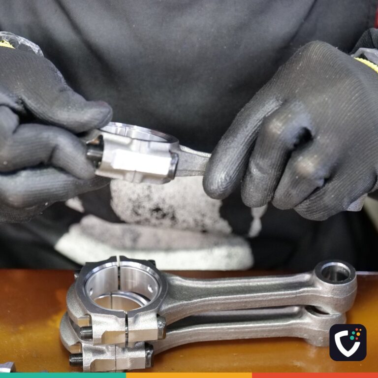 A technician wearing grip gloves checking a connecting rod bearing shell at a workbench, with a second assembled rod resting in front of him.
