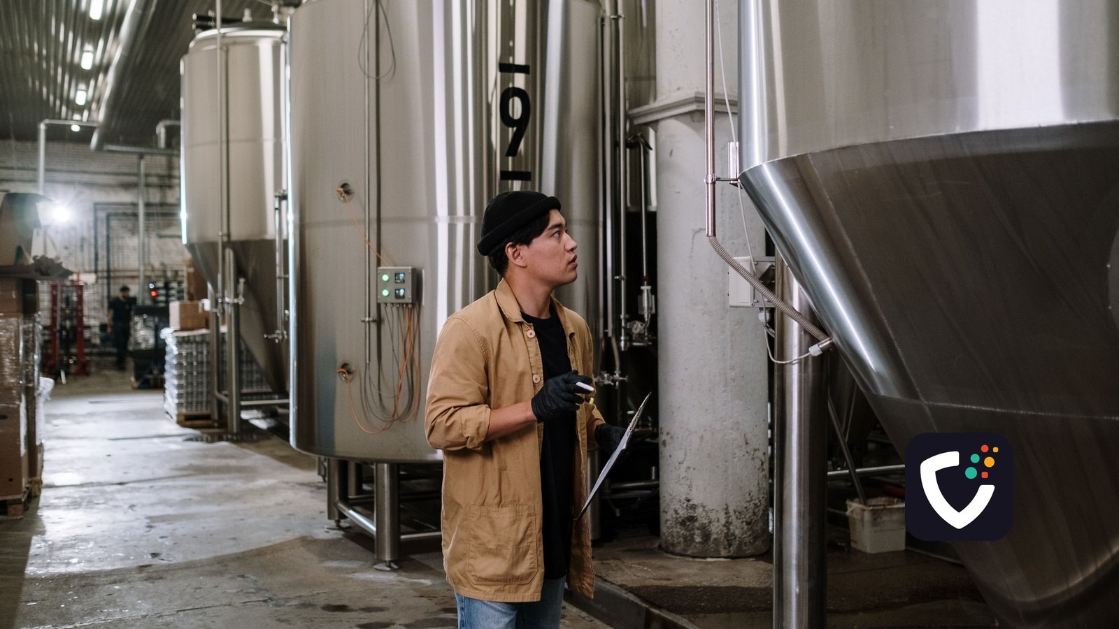 An inspector holding a clipboard, looking up at a row of stainless steel process tanks in a production hall.