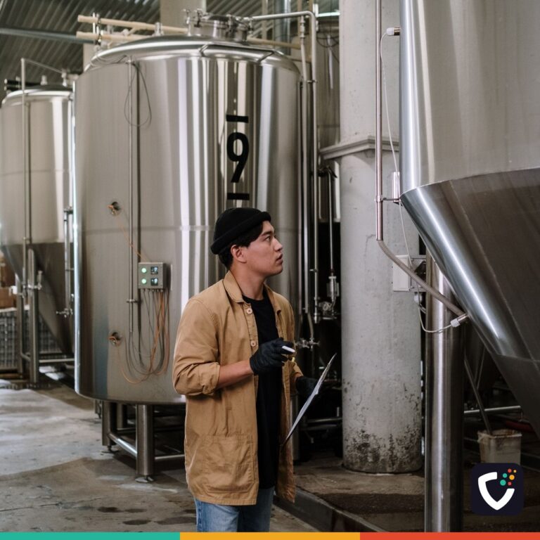 An inspector holding a clipboard, looking up at a row of stainless steel process tanks in a production hall.
