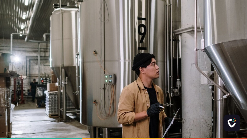 An inspector holding a clipboard, looking up at a row of stainless steel process tanks in a production hall.