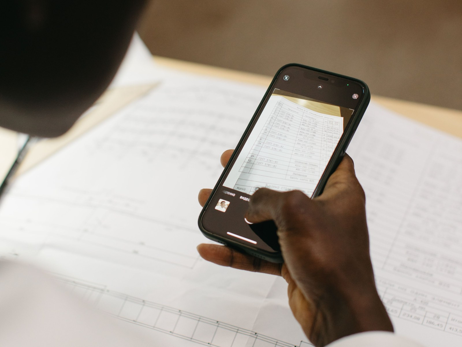 A hand holding a paper form flat on a desk while a phone held in the other hand photographs it.