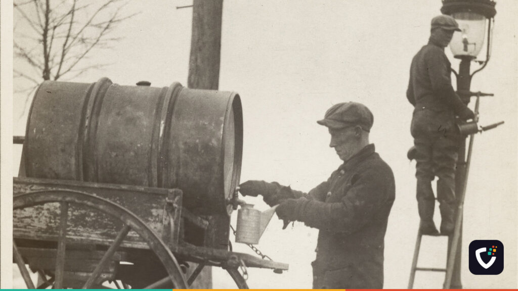 A black and white press photograph from 1926. A lamplighter in a flat cap fills a can from a tank on a horse cart, while a second man stands on a ladder at the top of a gas street lamp, servicing it.