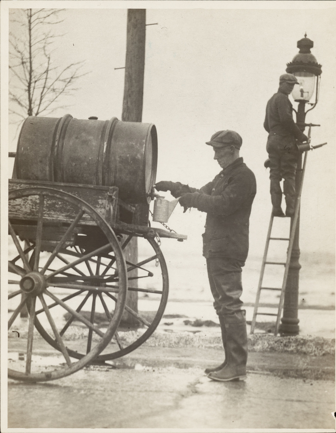A black and white press photograph from 1926. A lamplighter in a flat cap fills a can from a tank on a horse cart, while a second man stands on a ladder at the top of a gas street lamp, servicing it.