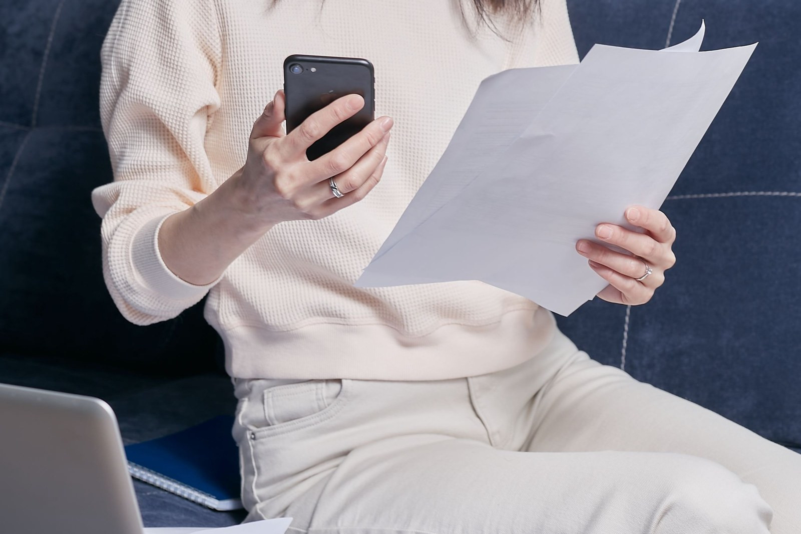 Somebody sitting on a sofa, holding a letter in one hand and a phone in the other, photographing it, with a laptop open beside them.