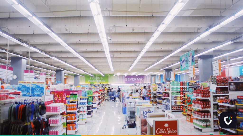 A wide supermarket aisle with lit ceiling strips, well stocked shelves and a few shoppers in the distance.