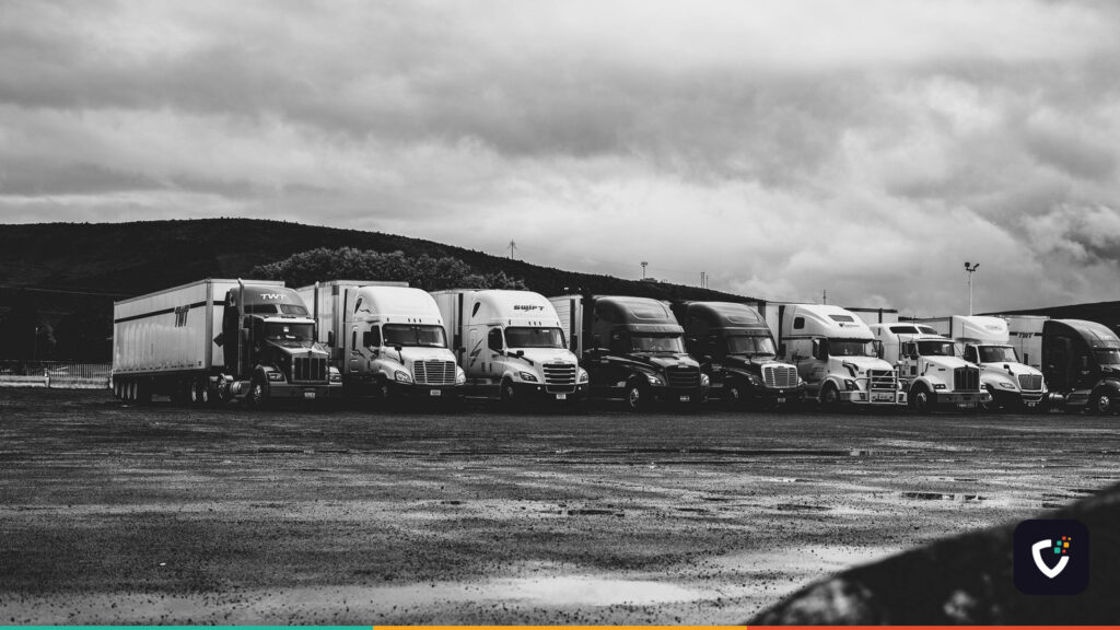 A row of parked American articulated trucks in a wet lot under heavy cloud.