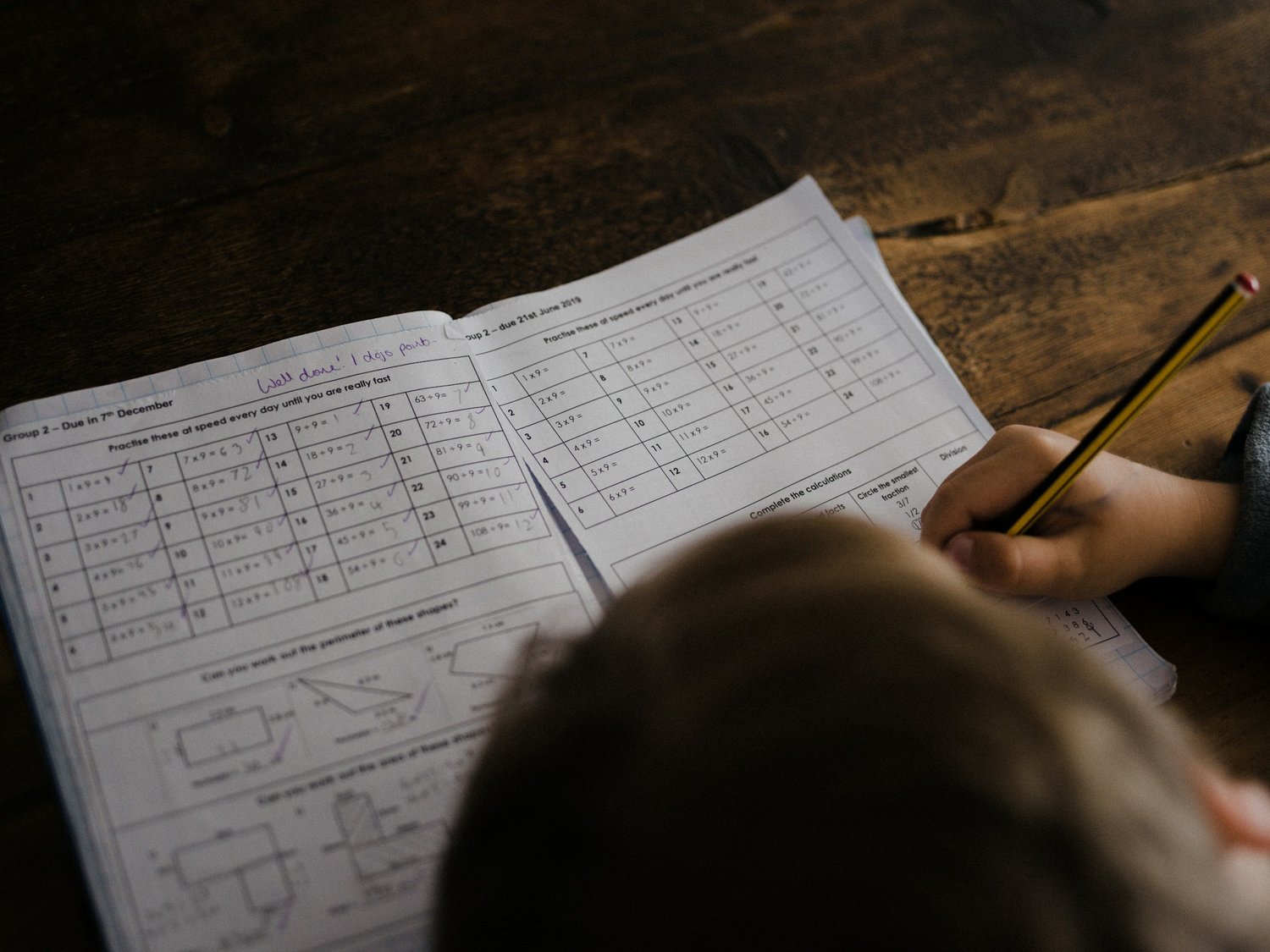 Over the shoulder of a child writing in a maths exercise book at a wooden desk.